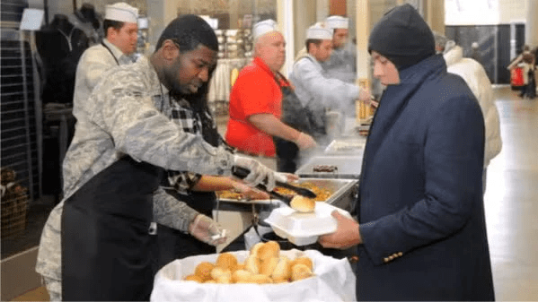 A volunteer serving food to a man in a winter coat at a community meal event, with others preparing food in the background.
