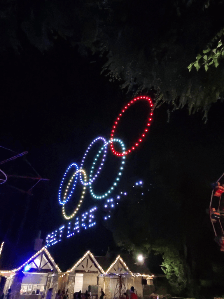 Drone show featuring colorful illuminated rings forming the Olympic symbol in the night sky above Liberty Park, with festive lights adorning the park buildings.