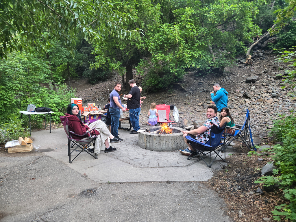 A group of people gathered around a bonfire at Ledgemere Picnic Area, enjoying food and conversation in a lush green setting.