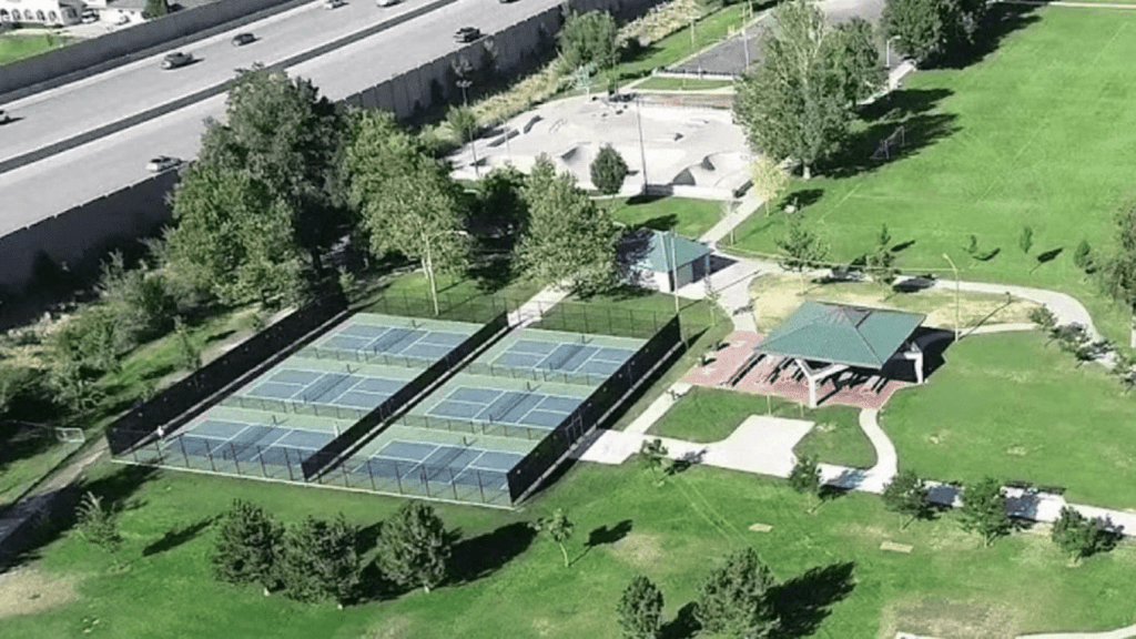 Aerial view of a park featuring multiple pickleball courts, green grass areas, trees, and a pavilion, with a nearby road visible.